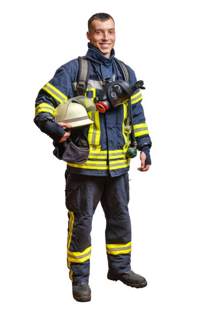 joven bombero sonriente con un uniforme ignífugo se para y mira a la cámara con un casco en las manos. - fire department heroes portrait occupation fotografías e imágenes de stock
