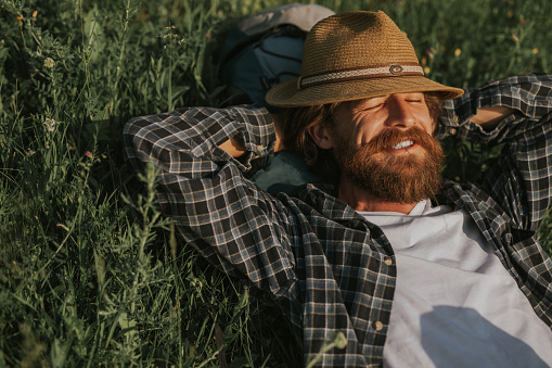 From above happy bearded man with closed eyes chewing straw and holding hands behind head, while resting on green grass during trip in countryside on sunny day