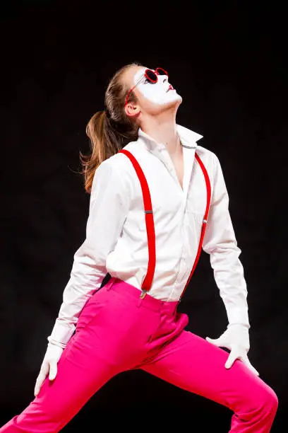 Photo of Portrait of male mime artist, isolated on black background. Man in suspenders and pink trousers is posing touching his stomach. Symbol of popular, dance, 90s