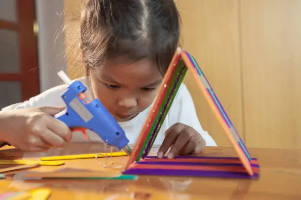 Photo of Asian child girl glueing colored ice cream sticks by hot melt electrical glue gun. Children have fun to make house on a handicraft project.