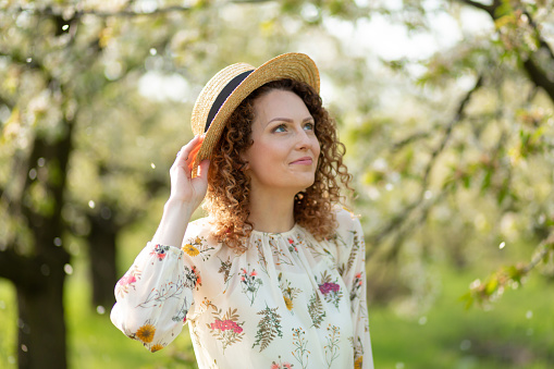 Portrait young attractive woman with curly hair in stylish wicker hat enjoys blooming green garden in spring day.