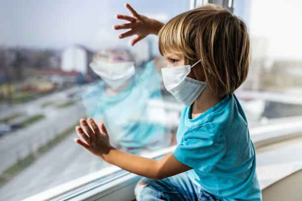istock Lonely little boy with face mask looking through window. 1216333353