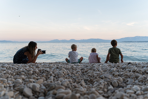 Young mother with professional dslr camera taking photos of her three kids, two boys and a girl, sitting on pebble beach in the evening.