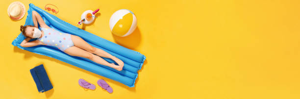 top view on a little girl in a swimsuit and a protective mask resting on an inflatable blue mattress on a yellow background, concept of summer vacation. banner copy space - bikini summer vacations looking down imagens e fotografias de stock