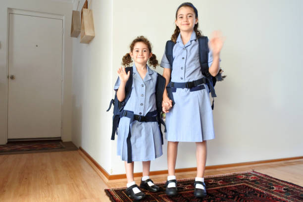 Two happy sisters (age 10 and 6) looking at camera and waving goodbye to parents before going to school together. Indoor home. Real people. Copy space