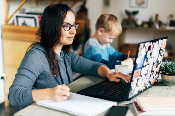 family education Concept of work from home and home family education. Mom and son are sitting at the desk. Business woman works on the Internet in a laptop, a child writes in a notebook. working at home with children stock pictures, royalty-free photos & images