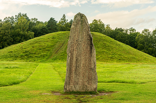 Close up view of a large rune stone standing at an old burial ground in Sweden with a large grassy burial mound in the background