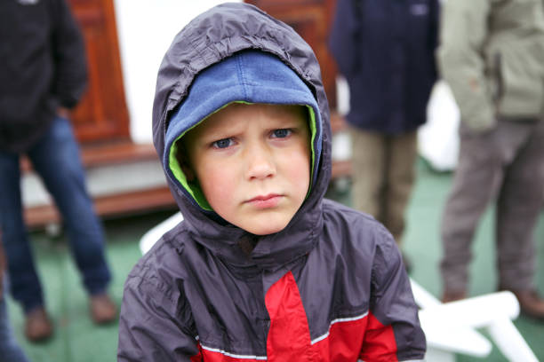Worried face on a young boy standing on a boat, staring at camera, being serious and concentrated, wearing rain jacket and hood on stock photo