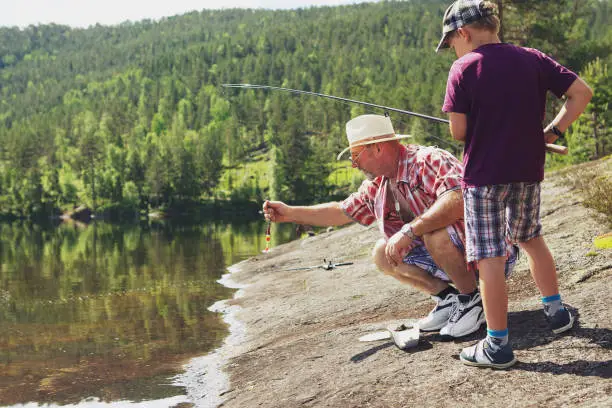 June 30, 2013 - Norway: grandfather teaching his grandson fishing - is helping with the fishing rod while relaxing on a rocky water's edge on the lake on a sunny day