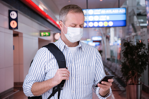 Man in protective mask standing in airport hall with smartphone and backpack, browsing, texting, using mobile app. Safe traveling.