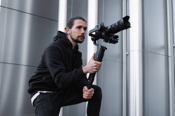 istock Young Professional videographer holding professional camera on 3-axis gimbal stabilizer. Pro equipment helps to make high quality video without shaking. Cameraman wearing white hoodie making a videos. 1216357905