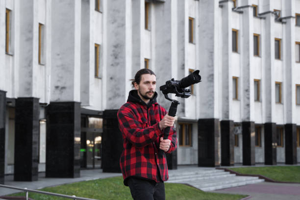istock Young Professional videographer holding professional camera on 3-axis gimbal stabilizer. Pro equipment helps to make high quality video without shaking. Cameraman wearing red shirt making a videos. 1216358026