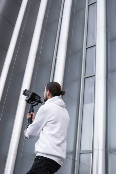 istock Young Professional videographer holding professional camera on 3-axis gimbal stabilizer. Pro equipment helps to make high quality video without shaking. Cameraman wearing white hoodie making a videos. 1216359159