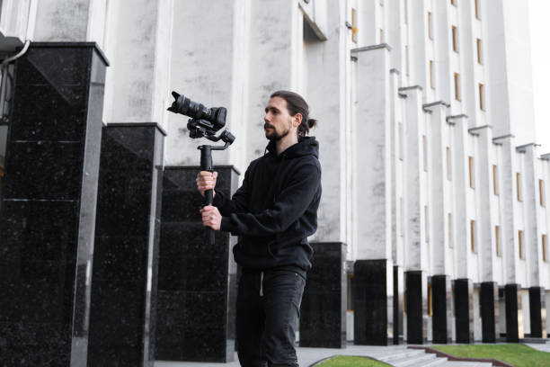 istock Young Professional videographer holding professional camera on 3-axis gimbal stabilizer. Pro equipment helps to make high quality video without shaking. Cameraman wearing black hoodie making a videos. 1216360542