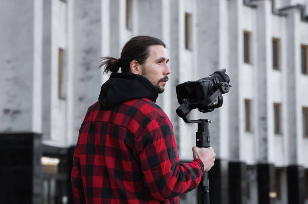 istock Young Professional videographer holding professional camera on 3-axis gimbal stabilizer. Pro equipment helps to make high quality video without shaking. Cameraman wearing red shirt making a videos. 1216361928