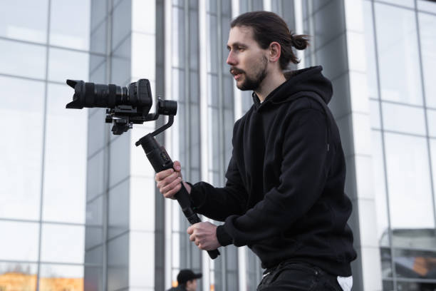 istock Young Professional videographer holding professional camera on 3-axis gimbal stabilizer. Pro equipment helps to make high quality video without shaking. Cameraman wearing white hoodie making a videos. 1216362533