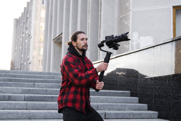 istock Young Professional videographer holding professional camera on 3-axis gimbal stabilizer. Pro equipment helps to make high quality video without shaking. Cameraman wearing red shirt making a videos. 1216363416