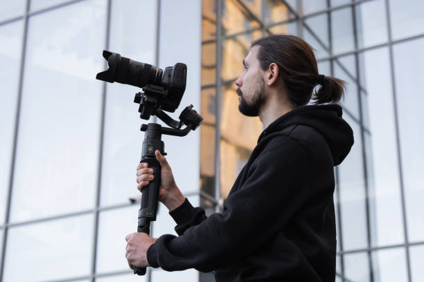istock Young Professional videographer holding professional camera on 3-axis gimbal stabilizer. Pro equipment helps to make high quality video without shaking. Cameraman wearing white hoodie making a videos. 1216363729