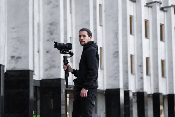 istock Young Professional videographer holding professional camera on 3-axis gimbal stabilizer. Pro equipment helps to make high quality video without shaking. Cameraman wearing black hoodie making a videos. 1216364147