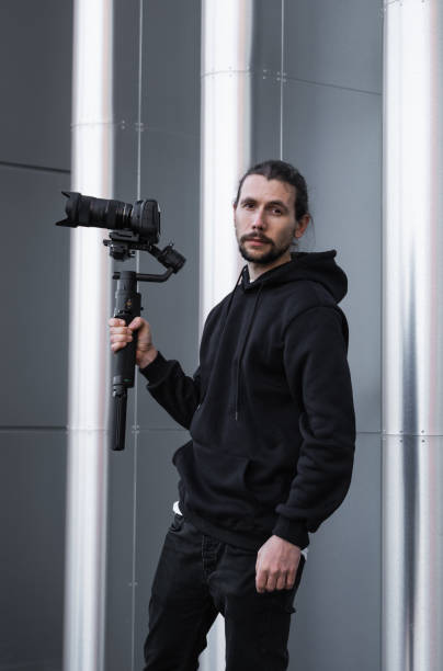 istock Young Professional videographer holding professional camera on 3-axis gimbal stabilizer. Pro equipment helps to make high quality video without shaking. Cameraman wearing white hoodie making a videos. 1216364985