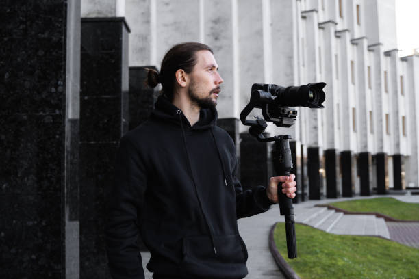 istock Young Professional videographer holding professional camera on 3-axis gimbal stabilizer. Pro equipment helps to make high quality video without shaking. Cameraman wearing black hoodie making a videos. 1216366040