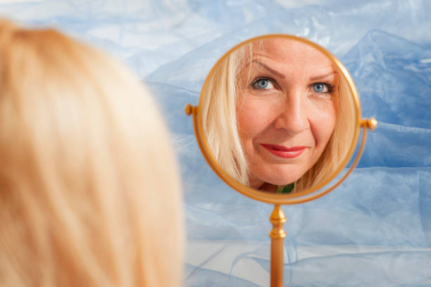 Reflection of the face of a middle-aged blonde woman in a round make-up mirror against a light blue wrinkled fabric background.