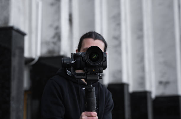 istock Young Professional videographer holding professional camera on 3-axis gimbal stabilizer. Pro equipment helps to make high quality video without shaking. Cameraman wearing black hoodie making a videos. 1216366318