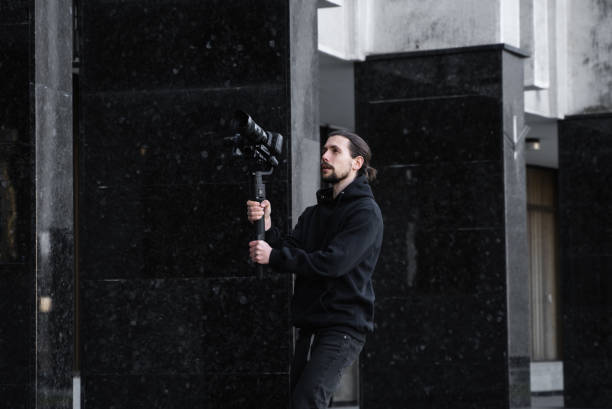 istock Young Professional videographer holding professional camera on 3-axis gimbal stabilizer. Pro equipment helps to make high quality video without shaking. Cameraman wearing black hoodie making a videos. 1216366515
