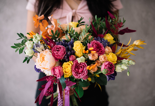 Very nice young woman holding big blossoming bouquet of campanella peach roses, misty bubbles roses, ranunculus, eustoma, pistachio fresh flowers in pink and yellow colors on the grey wall background