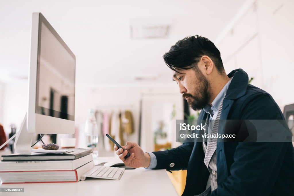 Man sitting behind the desk, looking at smartphone Young Asian Entrepreneur sitting behind the desk, using technology. Wasting Time Stock Photo