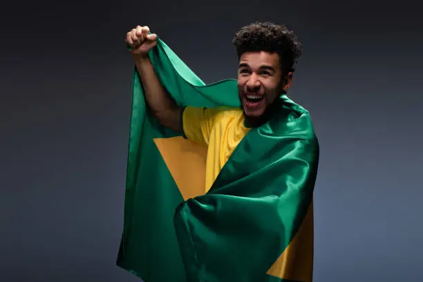 smiling african american football fan wrapped in brazilian flag on grey
