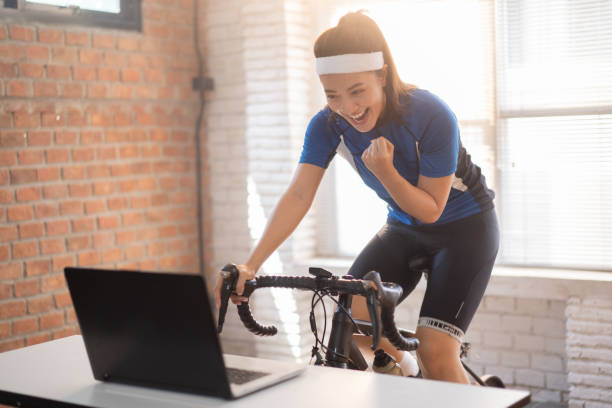 mulher asiática ciclista. ela está se exercitando em casa. ao pedalar no treinador e jogar jogos online de bicicleta, ela está feliz por ter sucesso. - short cycle - fotografias e filmes do acervo