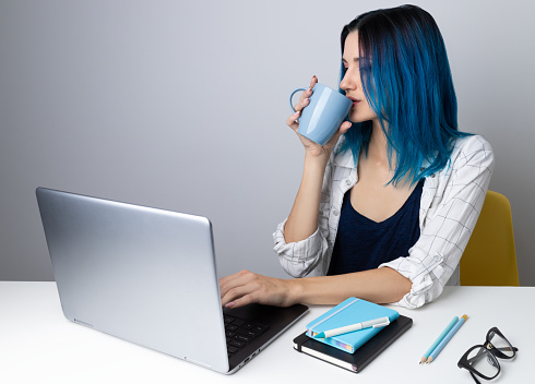 Happy young hipster woman with blue hair drink coffee or tea relax with laptop. Successful smiling girl working from home concept.