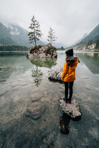 Rear view of a woman with a yellow jacket standing on rock in lake, Eibsee, Bavaria, Germany