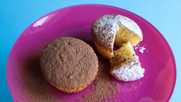 istock Two curd cakes sprinkled with chocolate and icing sugar on a pink plate, on a blue background. Dessert, a small cupcake. white baked cookies with an airy texture. Food concept 1216398803