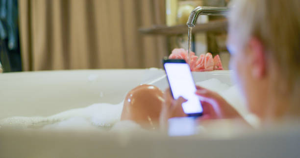 Woman using mobile phone in bathtub Over the shoulder view of a mature blond woman using a mobile phone while taking a bath in a bathtub primorska stock pictures, royalty-free photos & images