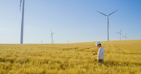 Side view shot of an engineer in a white helmet and a button down shirt standing in a wheat with wind turbines and using his mobile phone, Austria