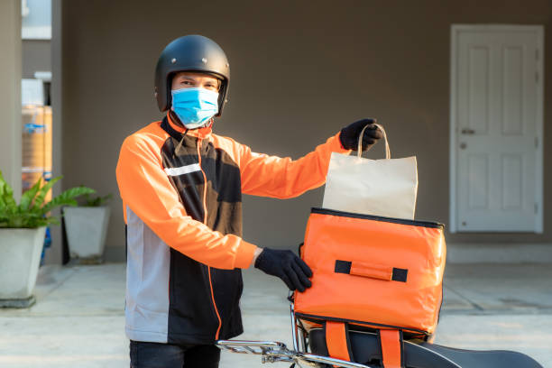 送貨亞洲男子穿著橙色制服的防護面具,準備在顧客家門前送送食品袋,用箱包在滑板車上,快遞食品配送和網上購物概念。 - 摄影 圖片 個照片及圖片檔