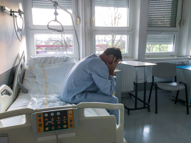 Worried patient sitting on bed in hospital with head in hands Side view of worried sad male adult patient with short hair sitting alone on a bed in a hospital room with head in hands hospital depression sadness bed stock pictures, royalty-free photos & images