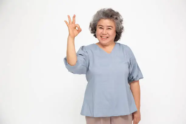 Photo of Portrait of happy senior asian woman gesture or showing hand ok and looking at camera isolated on white background, Older female feeling positive and enjoy concept
