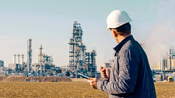 Waist up shot of an engineer in a white hardhat using a tablet with an oil refinery visible in the background.
