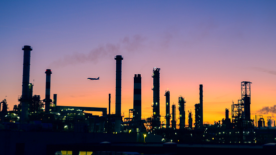 View of the silhouetted buildings of an oil refinery illuminated during sunset.