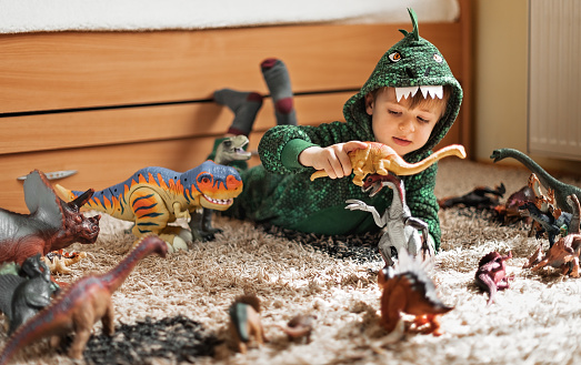 Adorable toddler boy in a dinosaur costume playing with his dinosaurs figures in his playroom
