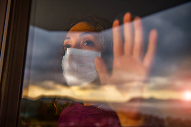 COVID 19 coronavirus quarantine self isolation social distancing Woman wearing a surgical mask and looking outside from her window. hopelessness photos stock pictures, royalty-free photos & images