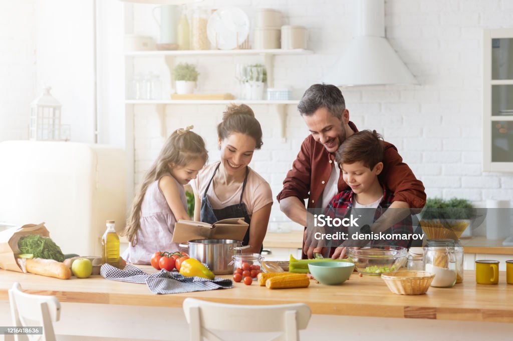 Happy family cooking together on home kitchen Happy family cooking together on kitchen. Mother and daughter reading recipe to father and son. Dad and boy chopping green vegetable leaf for salad. Home recreation and food preparation on weekend Family Stock Photo