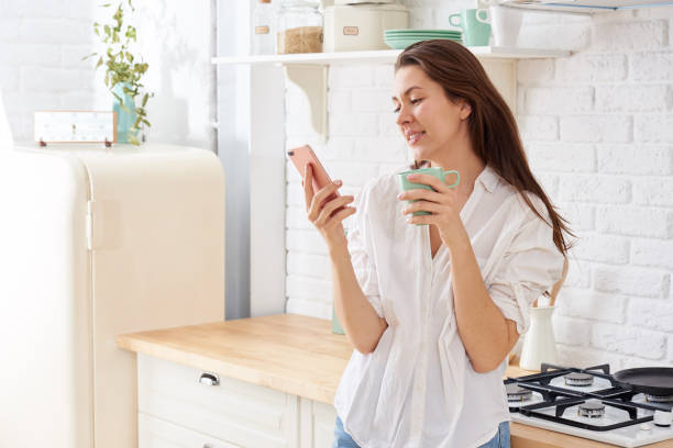 istock Young woman using smartphone leaning at kitchen table with coffee mug and organizer in a modern home. Smiling woman reading phone message. Brunette happy girl typing a text message 1216425208