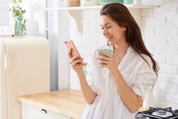 istock Young woman using smartphone leaning at kitchen table with coffee mug and organizer in a modern home. Smiling woman reading phone message. Brunette happy girl typing a text message 1216425474