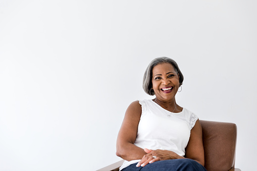 An attractive senior woman laughs cheerfully while smiling at the camera. She is sitting in a comfortable chair in her home.