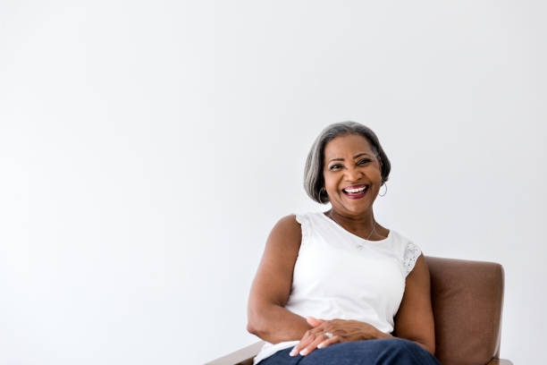 Portrait of beautiful senior woman An attractive senior woman laughs cheerfully while smiling at the camera. She is sitting in a comfortable chair in her home. woman sitting cross legged in chair stock pictures, royalty-free photos & images