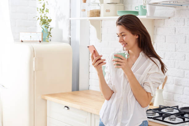 istock Young woman using smartphone leaning at kitchen table with coffee mug and organizer in a modern home. Smiling woman reading phone message. Brunette happy girl typing a text message 1216426502